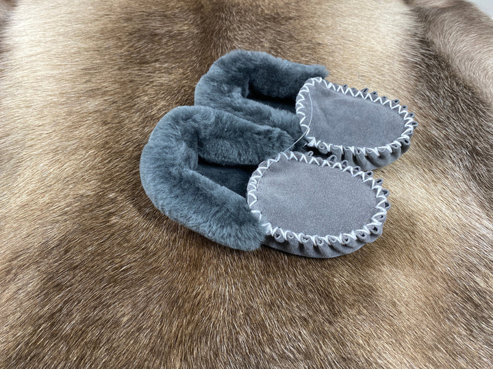 The Skin Thing grey sheepskin moccasin slippers with white stitching and EVA sole, displayed on a fur rug.