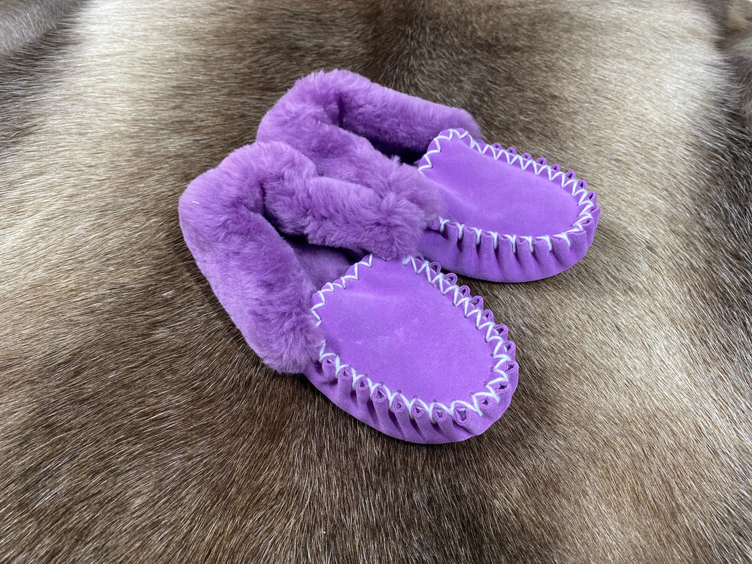 The Skin Thing purple sheepskin moccasin slippers with white stitching and fluffy lining on a fur rug.