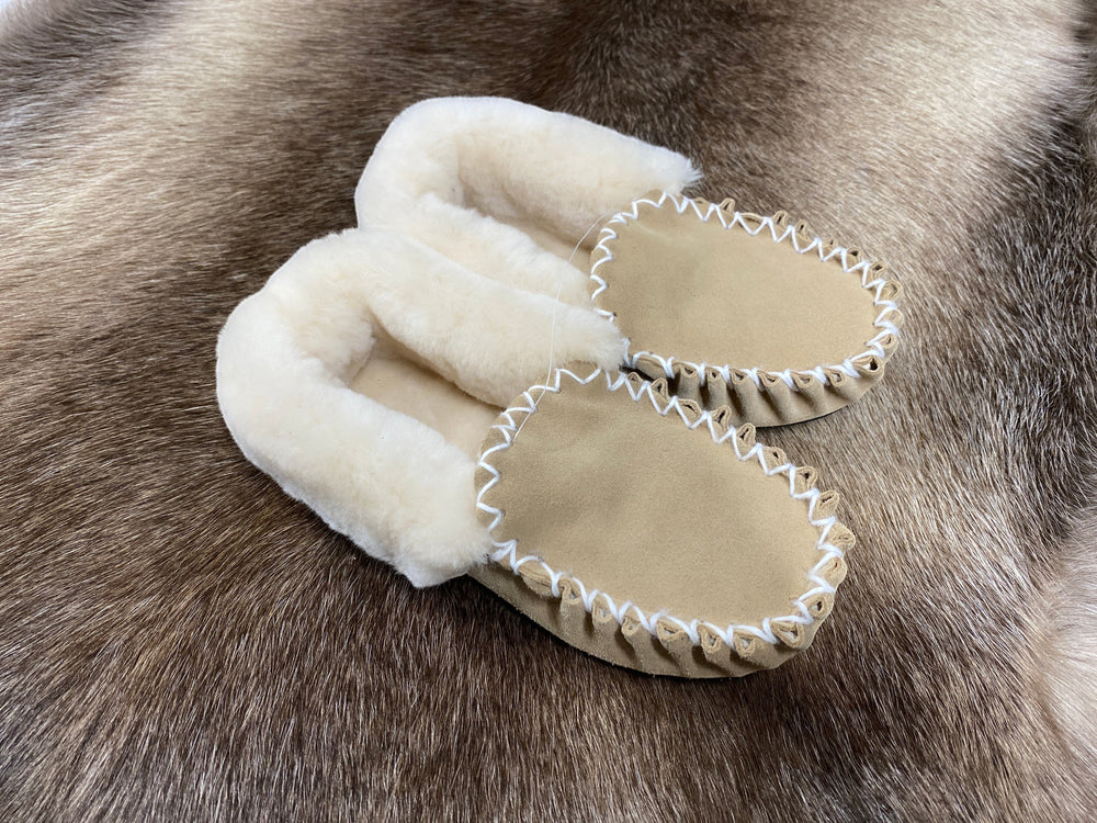 The Skin Thing tan sheepskin moccasin slippers with white stitching and fluffy lining, resting on a fur rug.