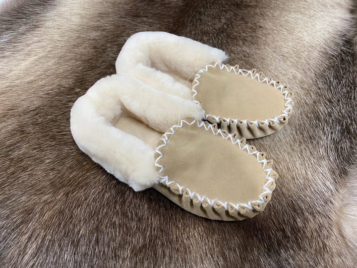 The Skin Thing tan sheepskin moccasin slippers with white stitching and fluffy lining, resting on a fur rug.